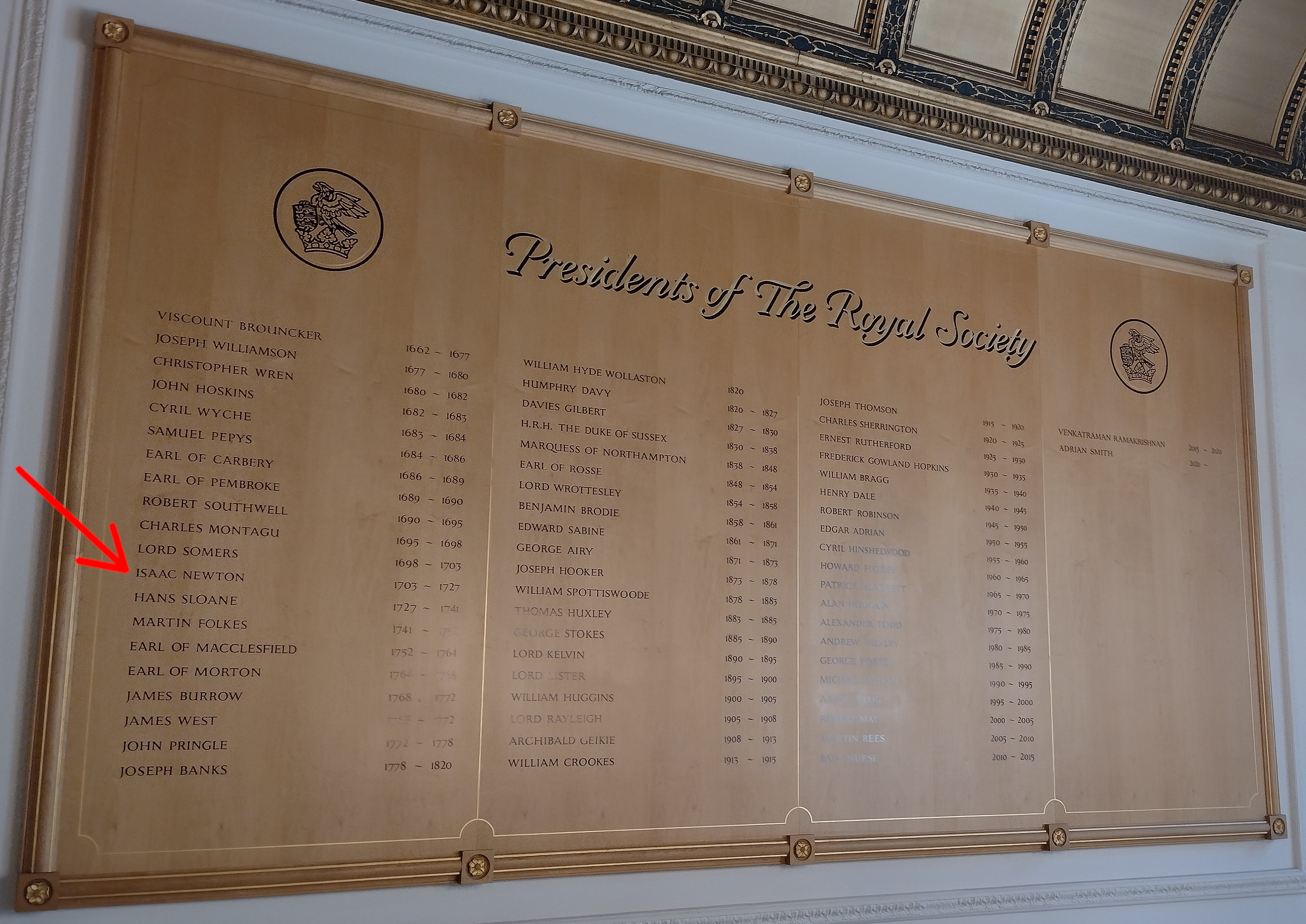 A large wooden plaque entitled 'Presidents of The Royal Society'. A red arrow points out Isaac Newton's name. The plaque lists the years of his presidency as 1703 – 1727.